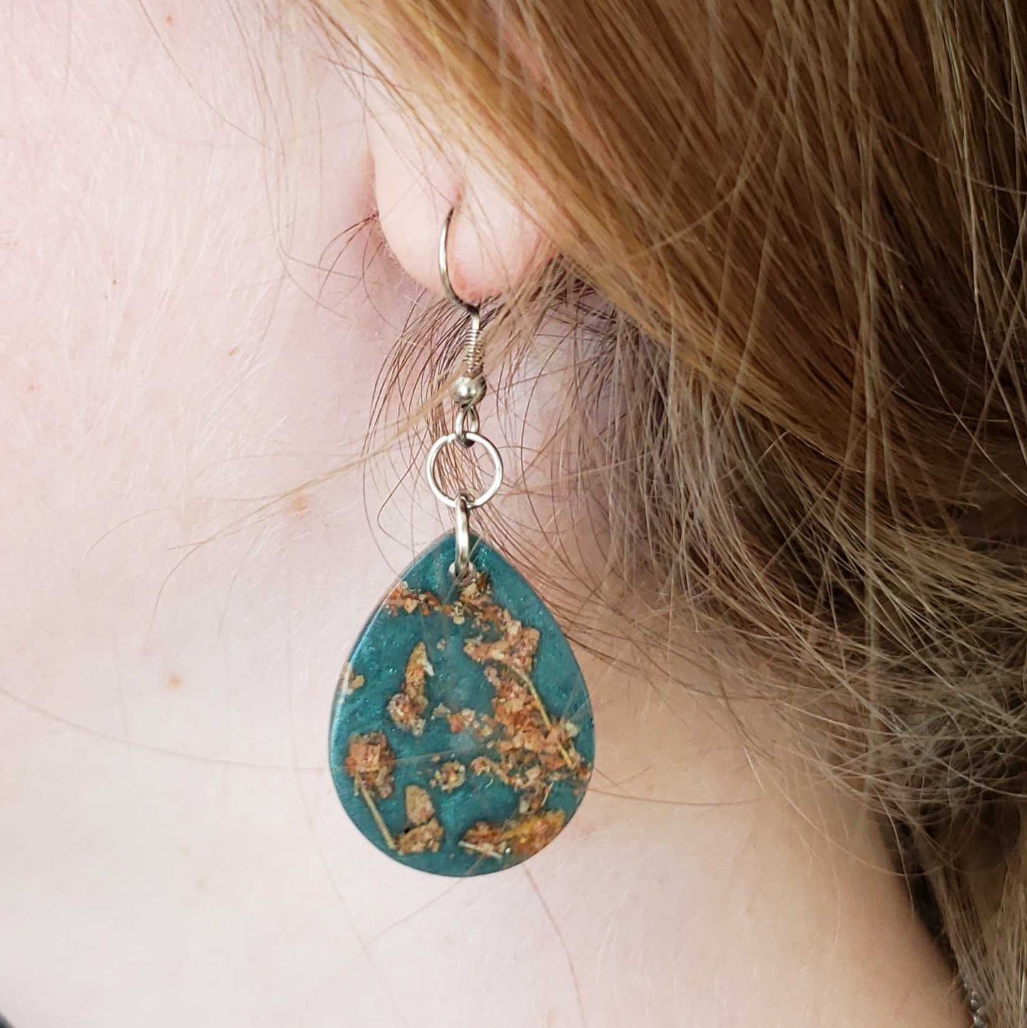 Teardrop-shaped turquoise earring with gold accents made from dried leaves worn by a person.