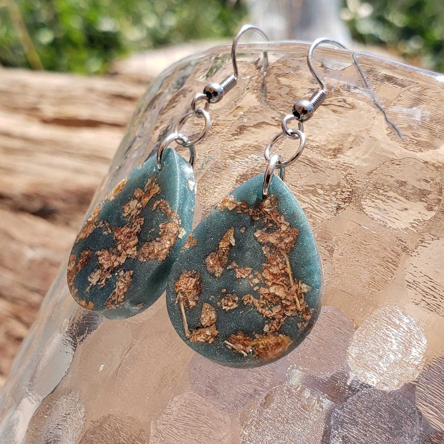 Teal dangle earrings with gold speckles made from dried leaves on a clear stone surface