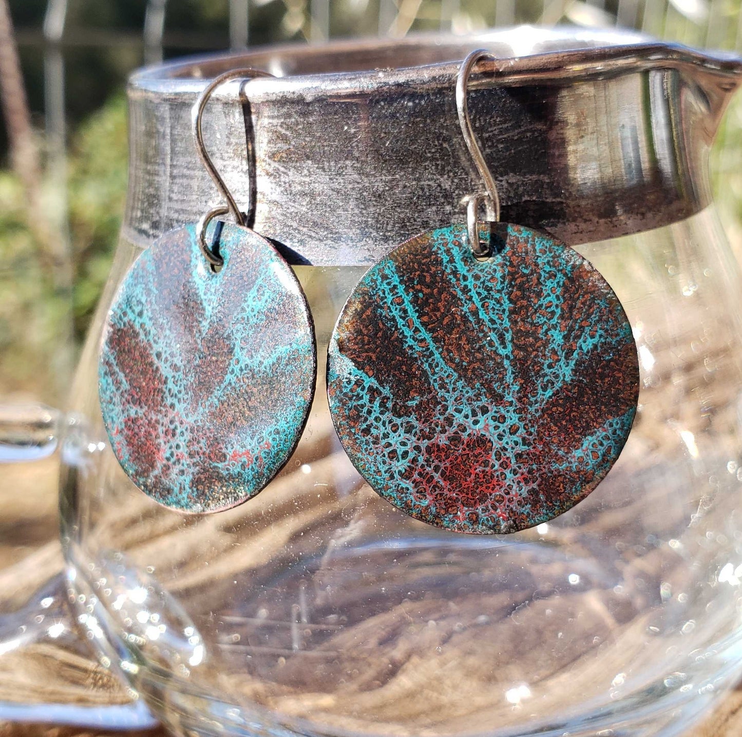 Enameled copper boho earrings with speckled design hanging from a glass container outdoors