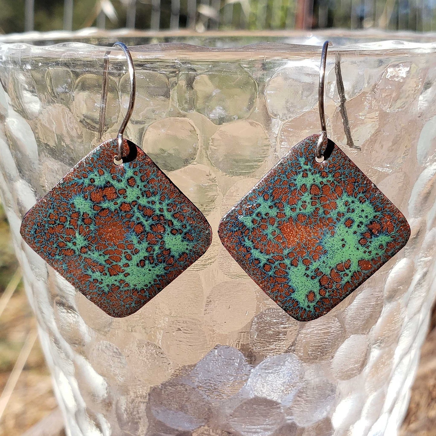 Square patterned boho copper enamel earrings on a textured surface with blurred natural background