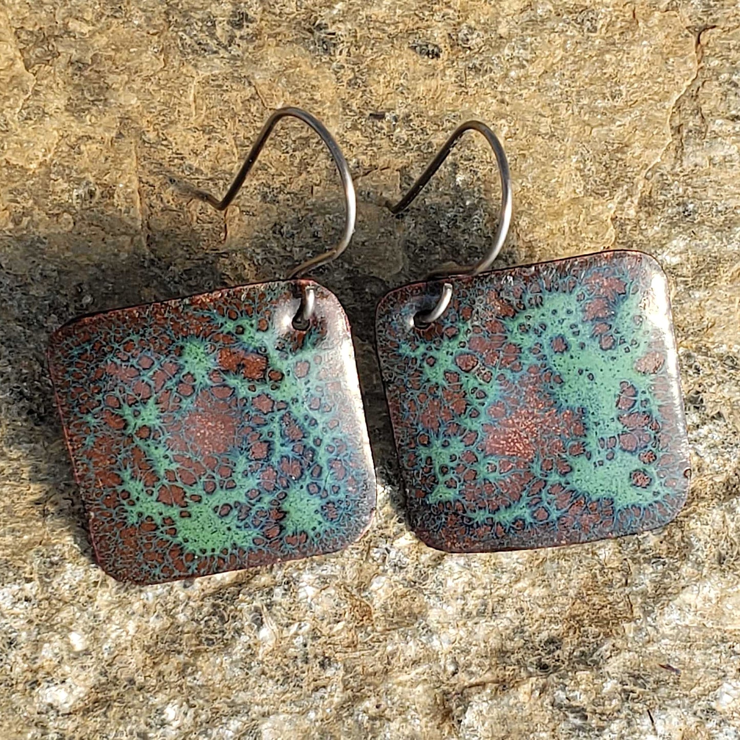 Pair of square copper enamel earrings with a textured, multicolored surface on a stone background
