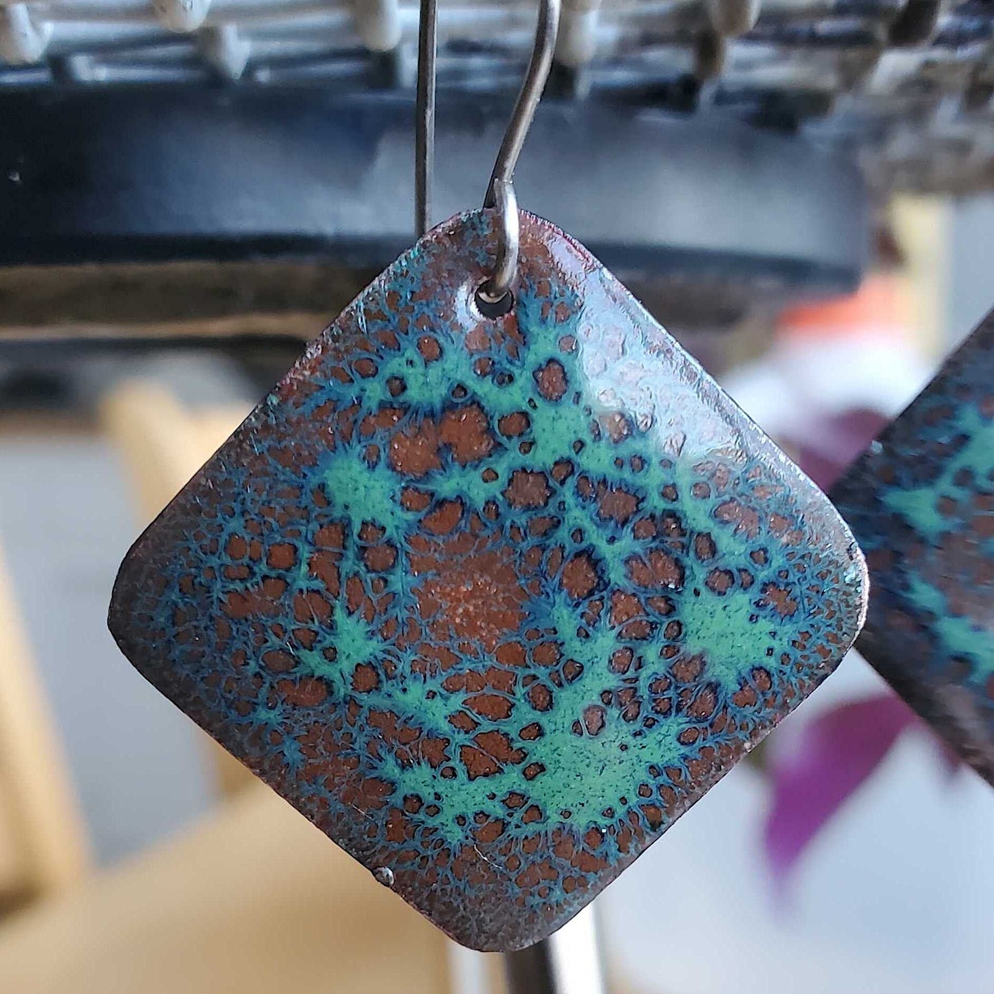 Square-shaped copper earrings with a copper and green speckled pattern on a blurred background.