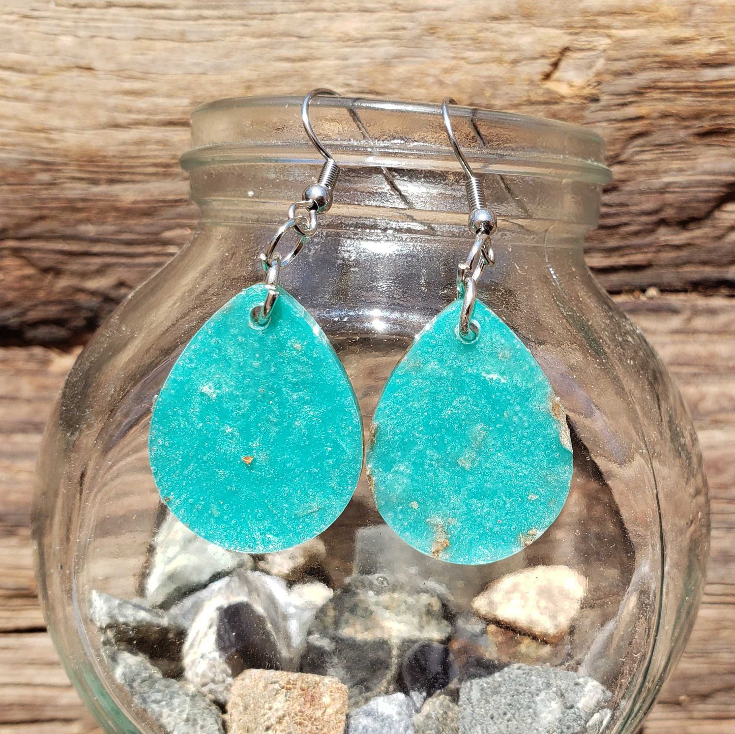 Back of teal teardrop earrings hanging from a glass jar with pebbles and stones.