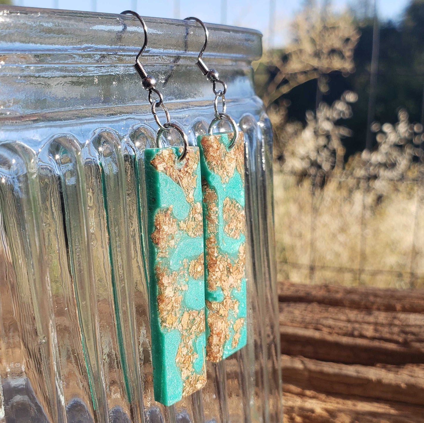 Turquoise earrings with gold accents made from dried leaves hanging on a glass jar outdoors.