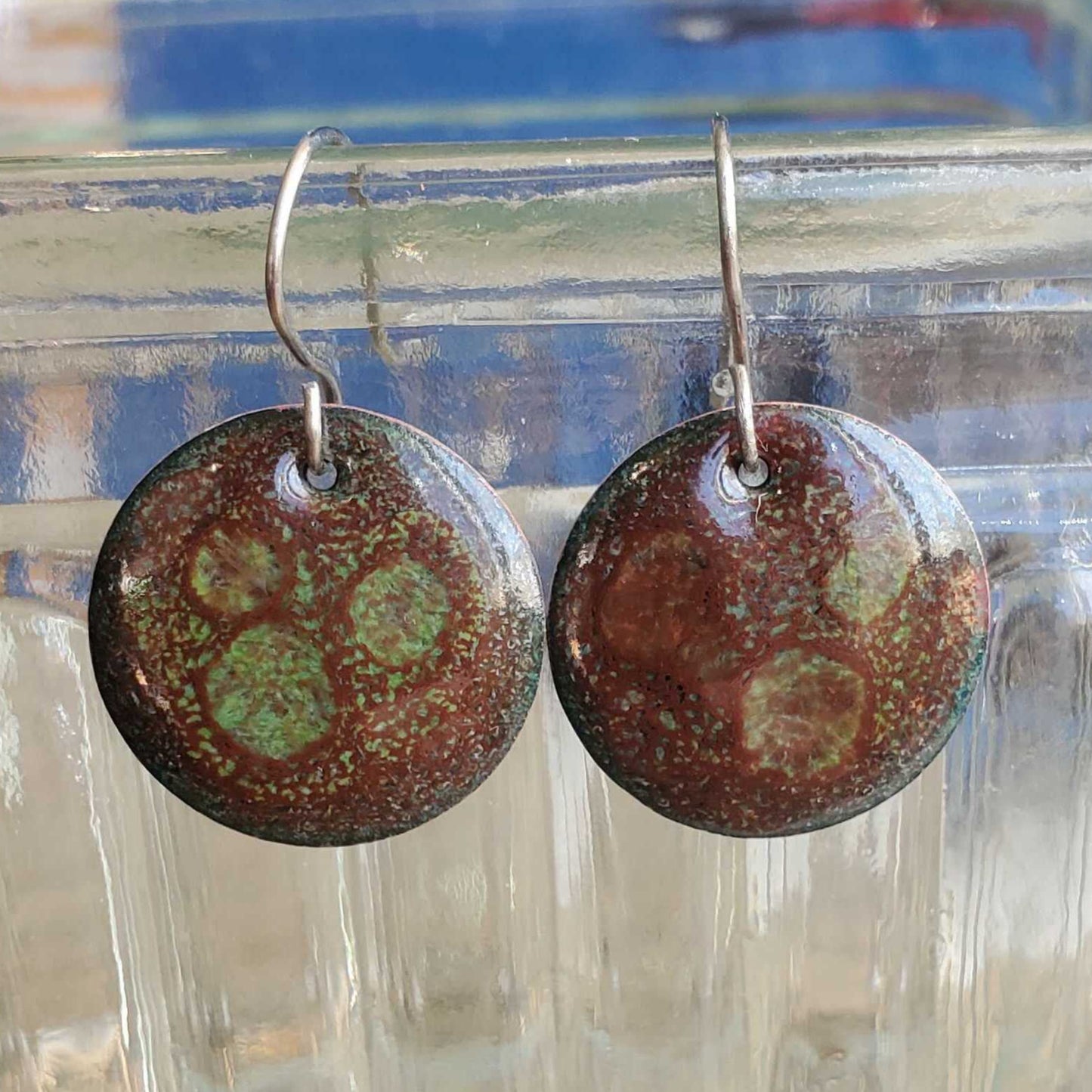Round copper enamel earrings with a brown and green pattern on a clear glass surface.