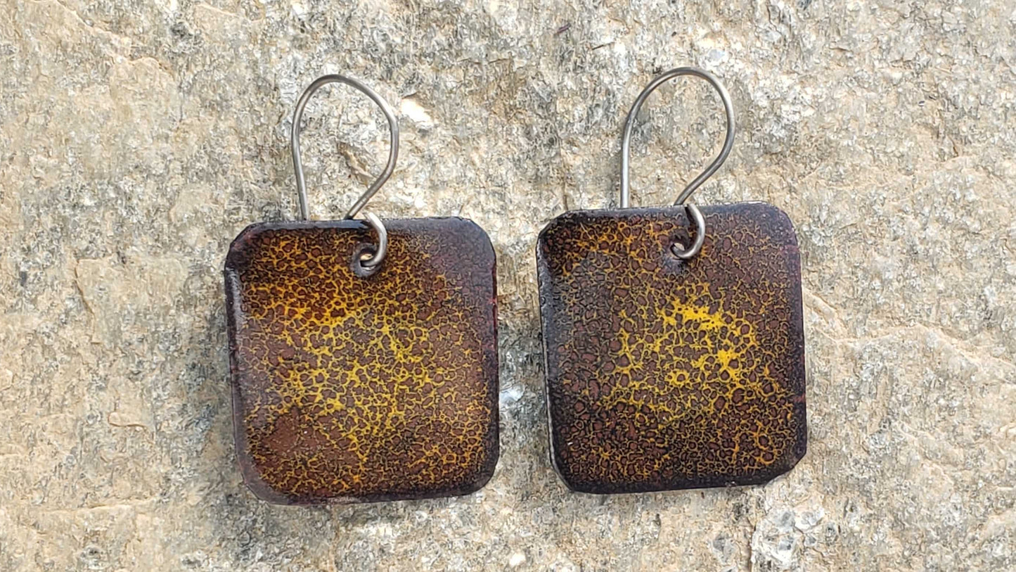 Pair of square copper enamel earrings with a brown rust-like finish on a stone surface
