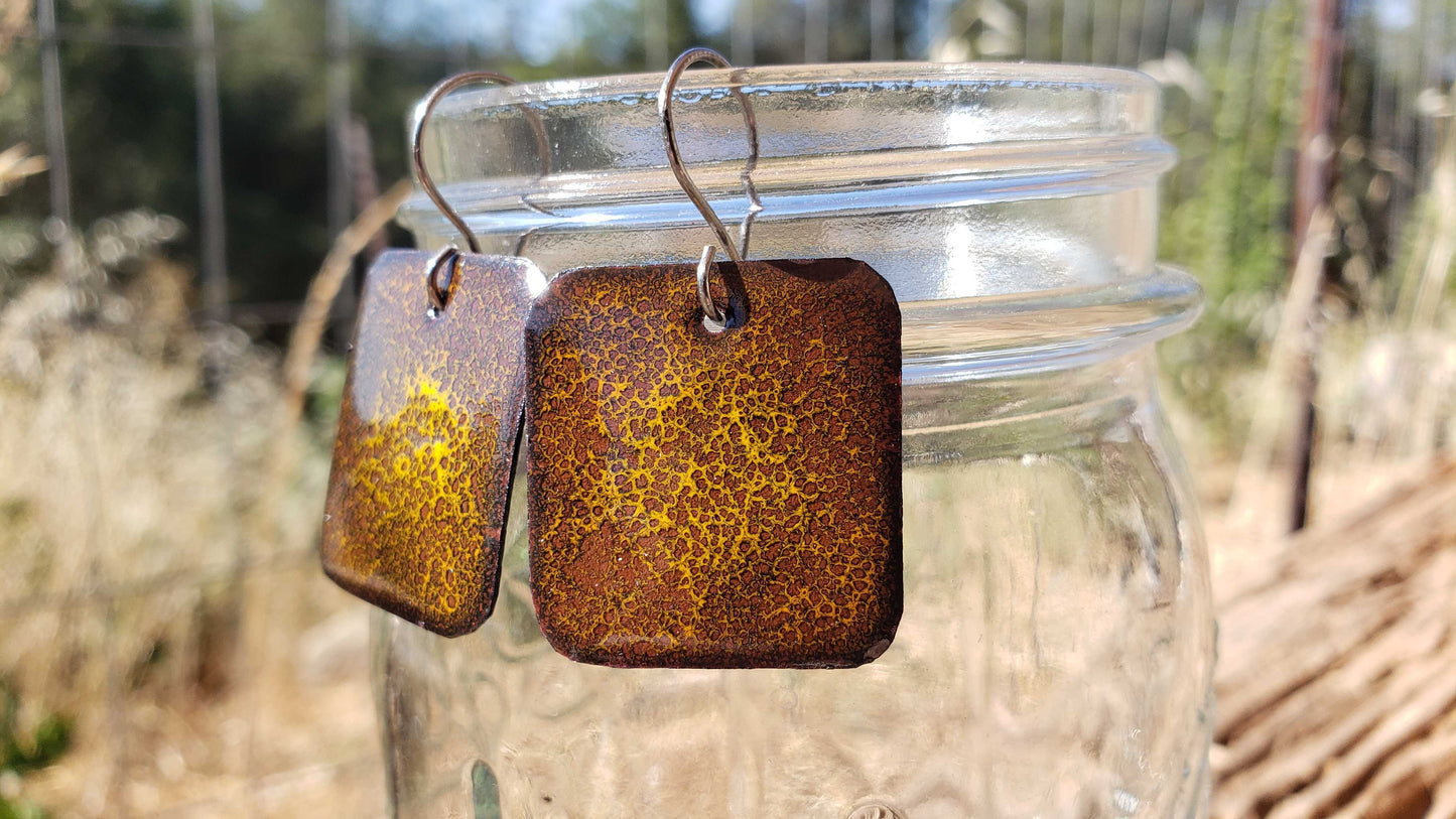 Brown square copper enamel earrings with a textured boho cheetah pattern hanging from a jar outdoors.