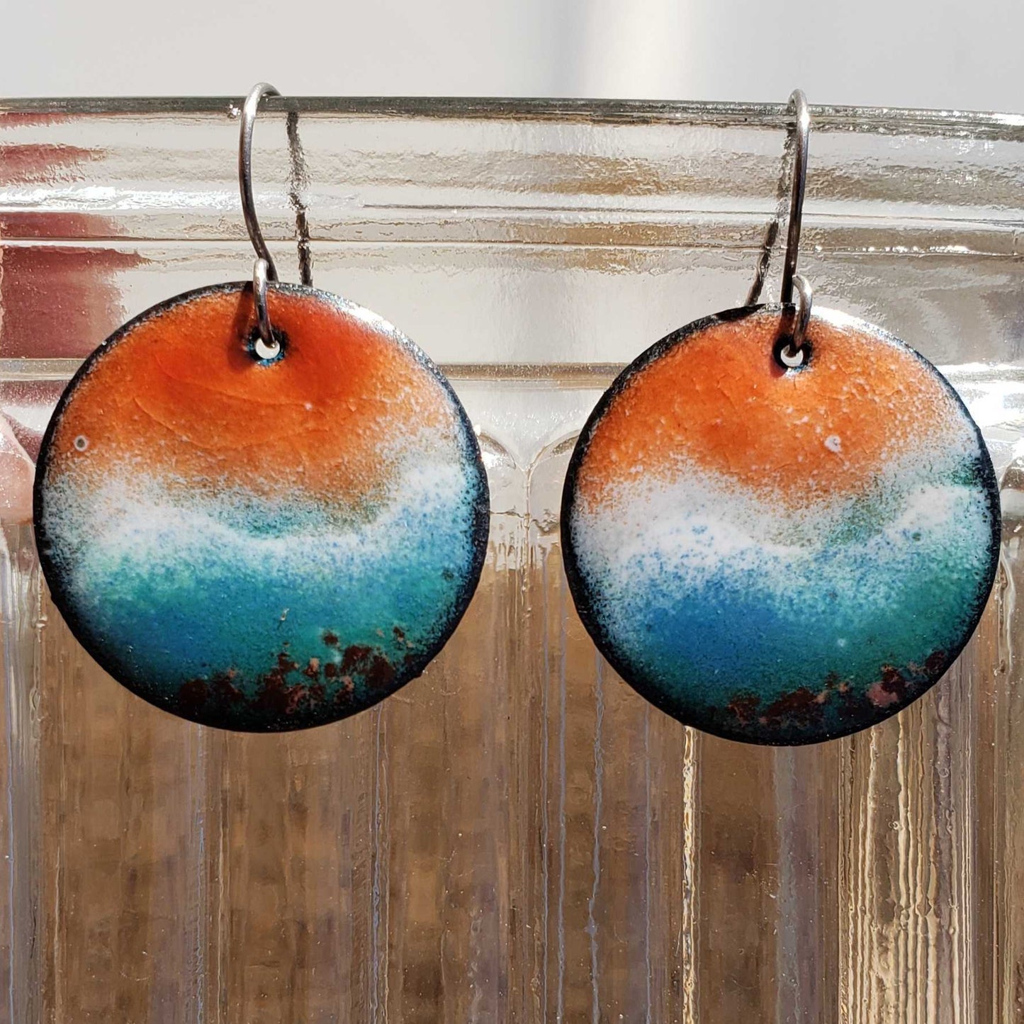 Round beach-inspired earrings with orange, blue, and white design hanging on a clear stand.