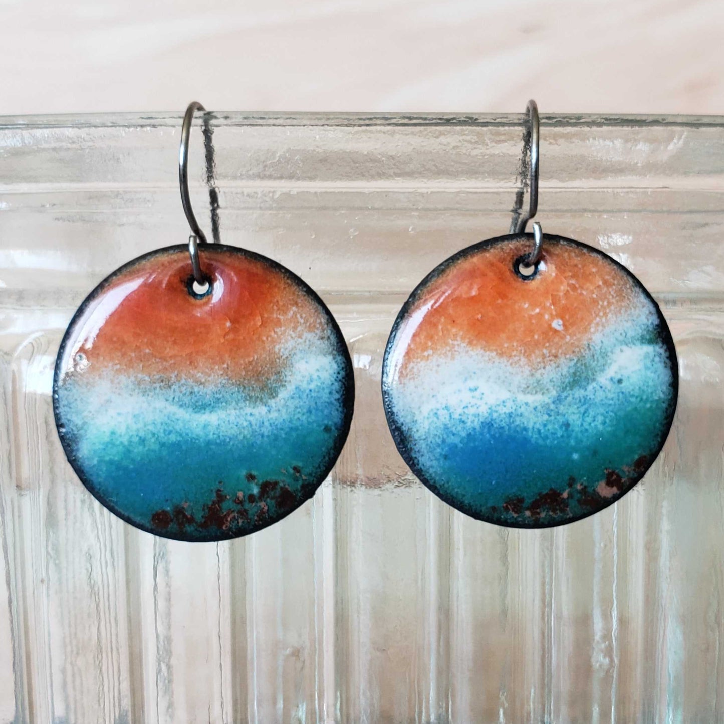 Round copper earrings with a blue and orange ocean scene gradient hanging from a glass jar.