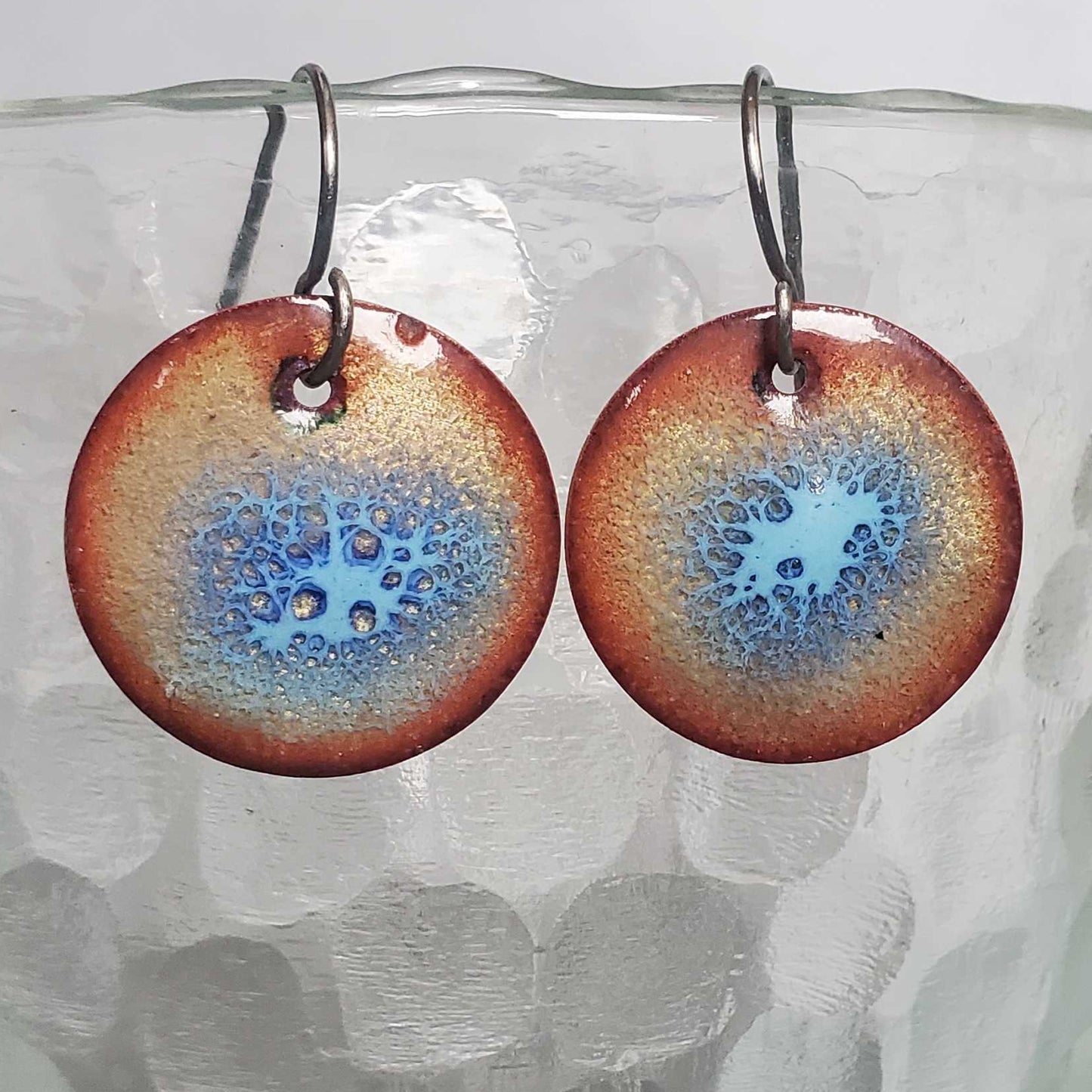 Round copper earrings with a blue and golden color scheme on a textured glass surface.