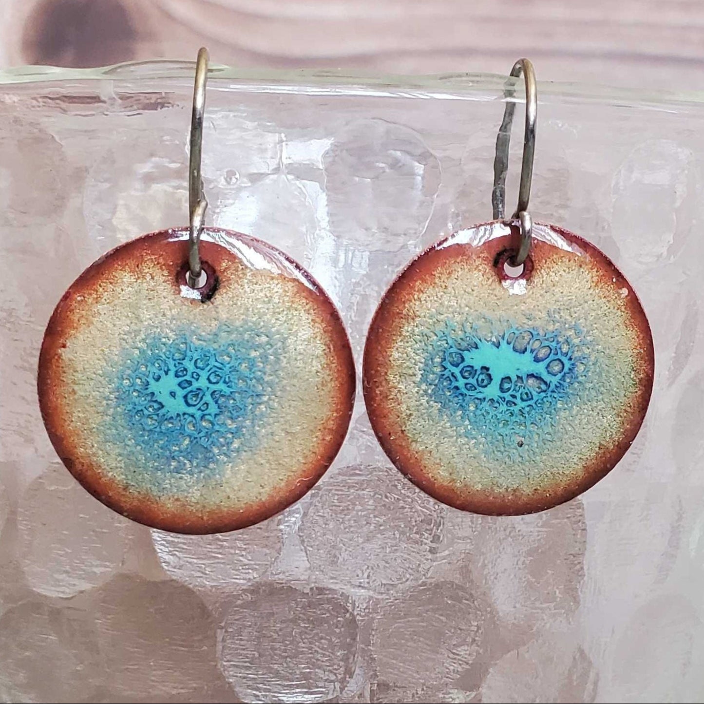 Round copper earrings with a blue and brown design on a glass surface.