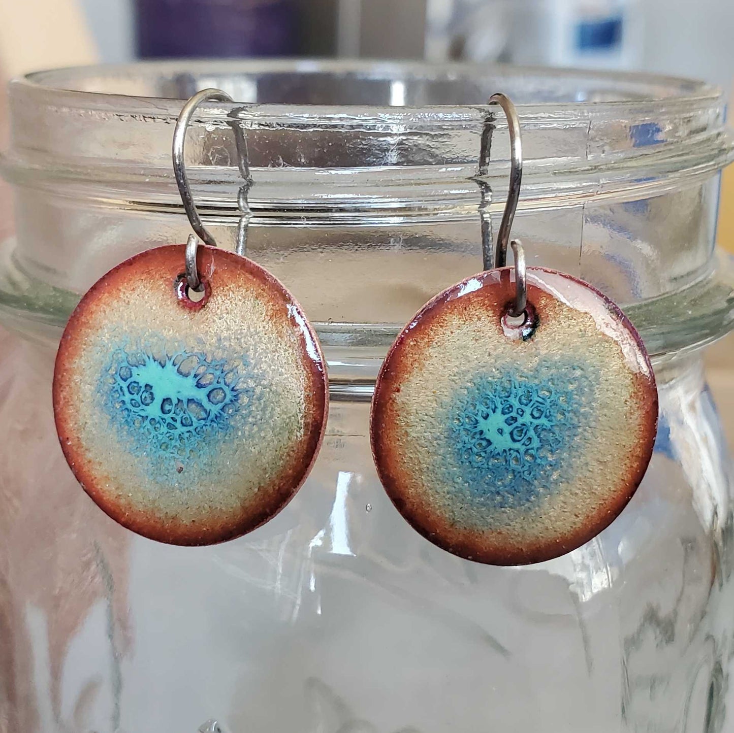 Round copper earrings with blue crackle design hanging from a glass jar on a wooden surface.