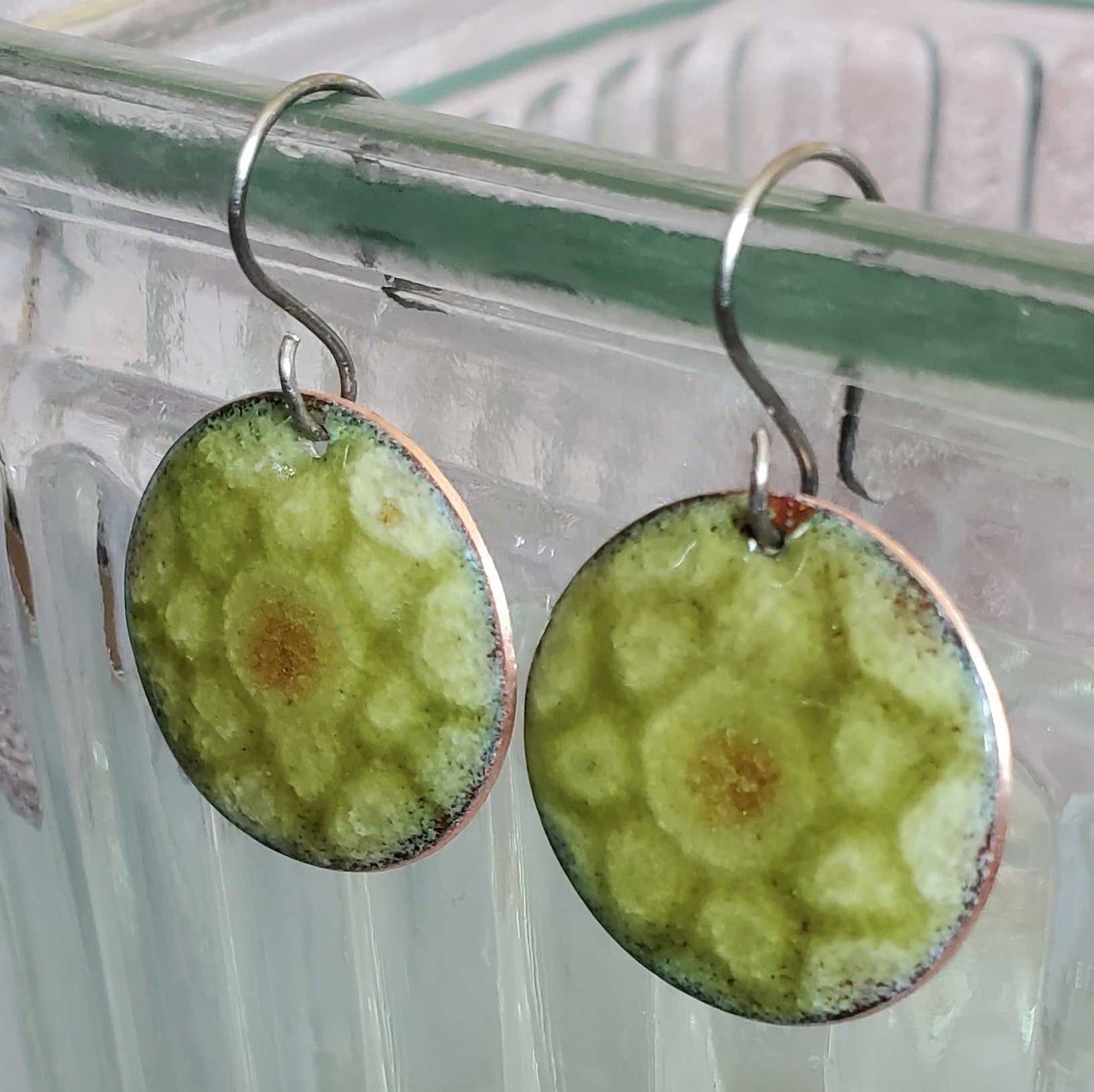 Round copper earrings with green and brown pattern hanging on a clear stand.