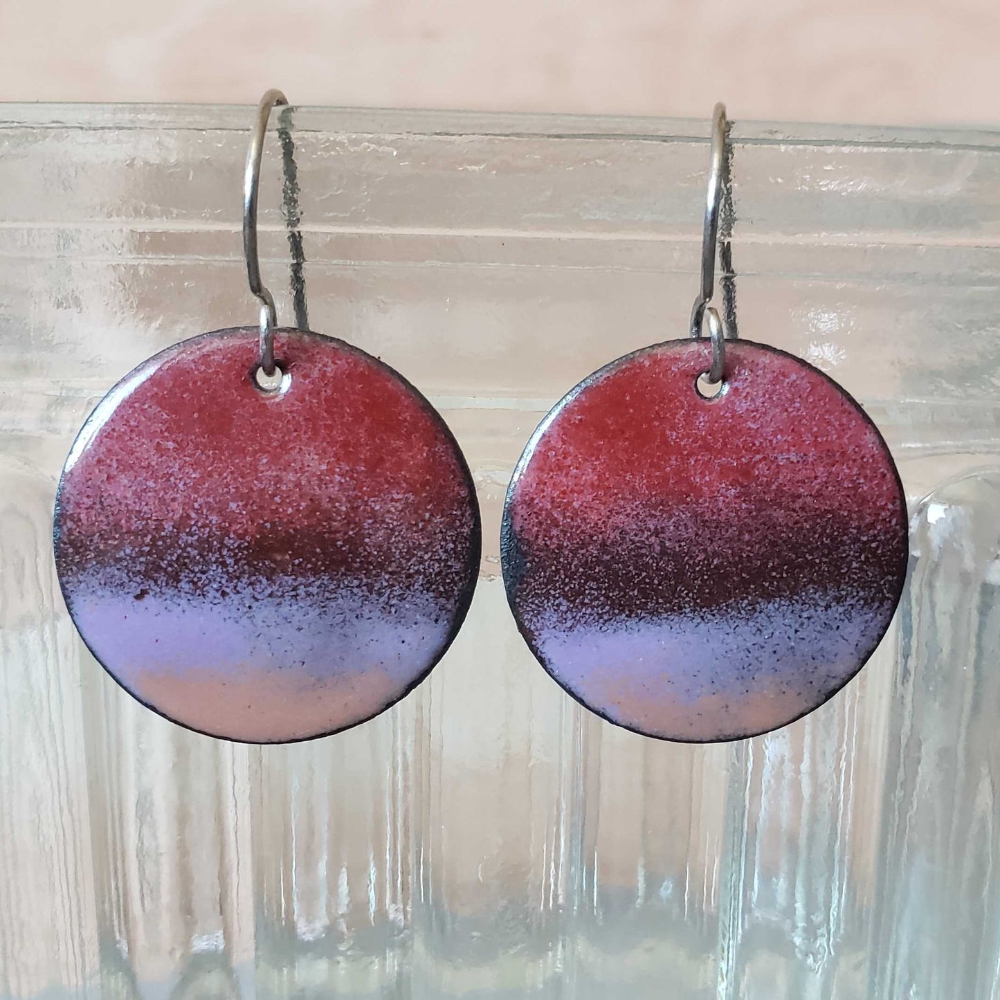 Round copper earrings with a gradient design in red and purple hanging on a clear glass vase.