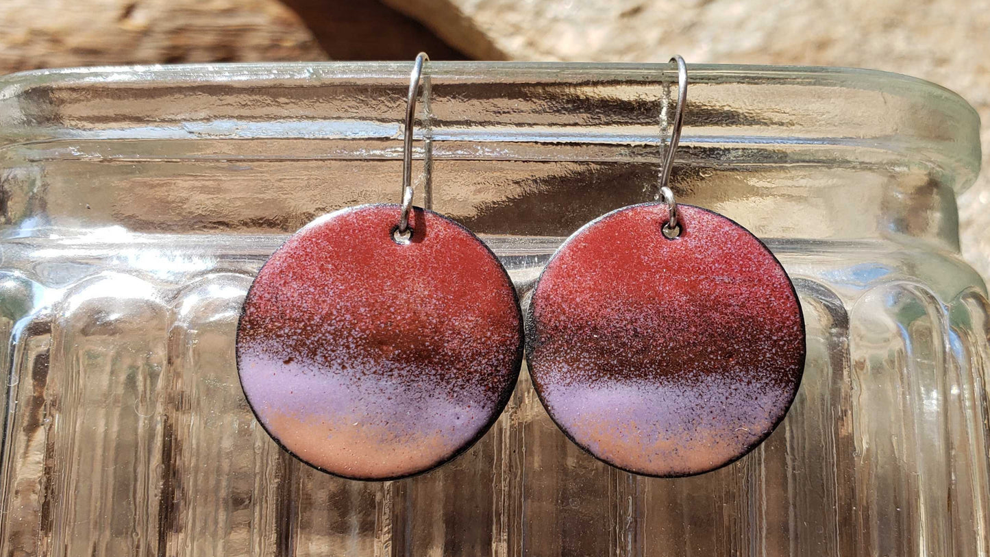 Round copper earrings with a gradient design hanging from a glass jar against a wooden background