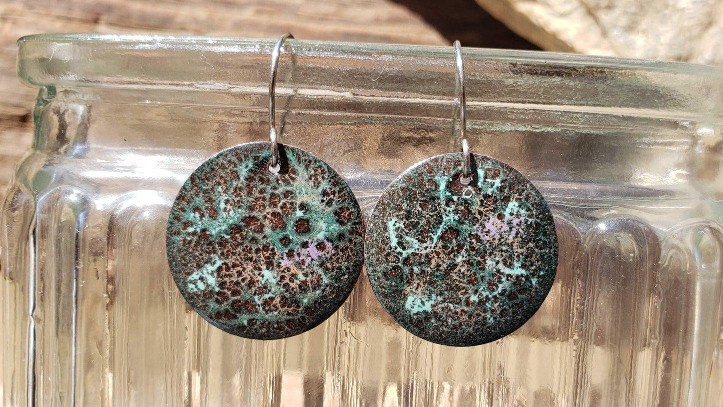 Round copper earrings with a brown crackle pattern hanging from a glass jar