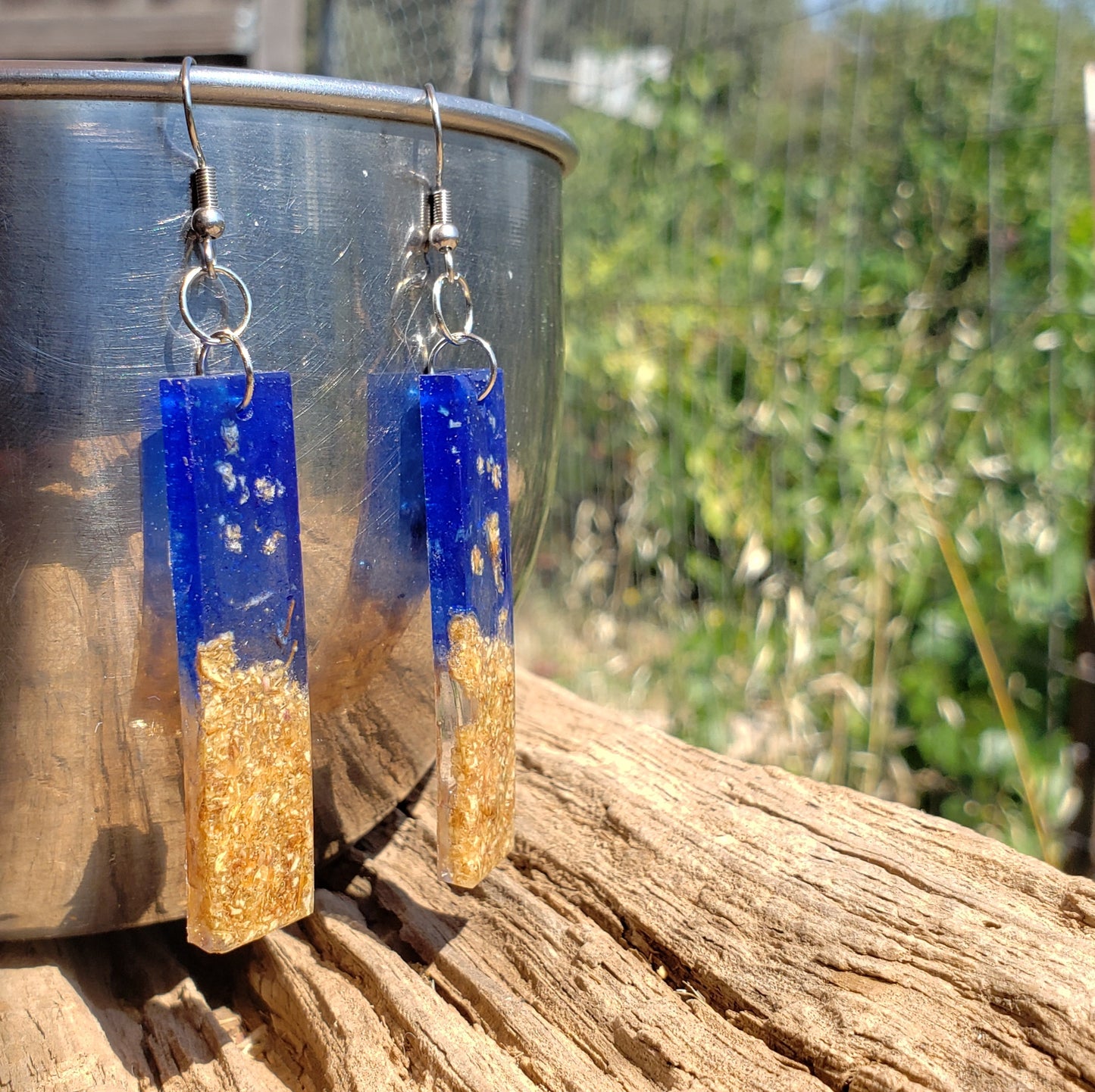 Blue and gold rectangular boho earrings with dried flowers hanging on a metal surface