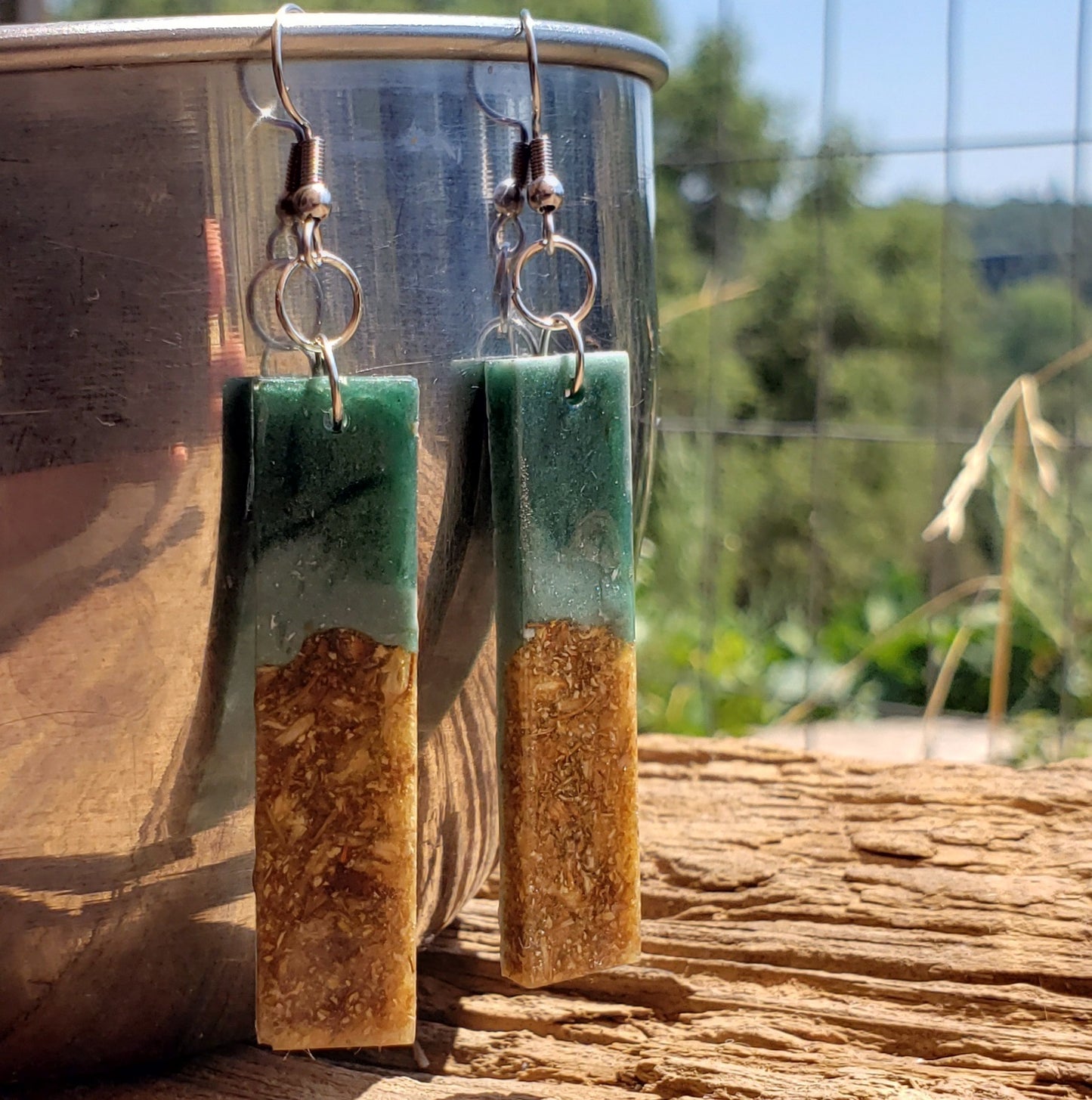 Green and brown rectangular earrings hanging from a metallic container on a wooden surface with a blurred natural background.