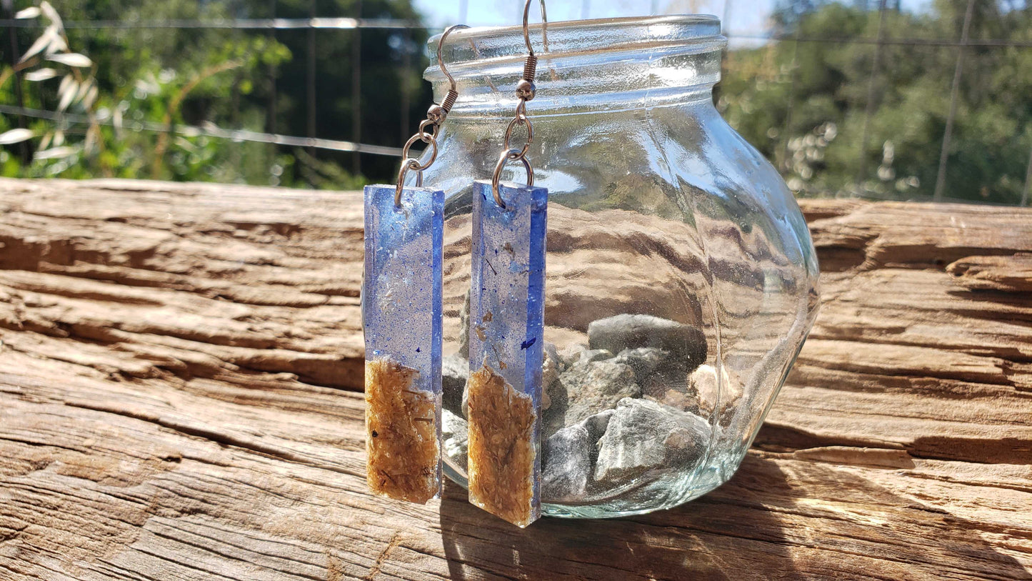 Pair of earrings with blue and brown elements hanging from a glass jar on a wooden surface.
