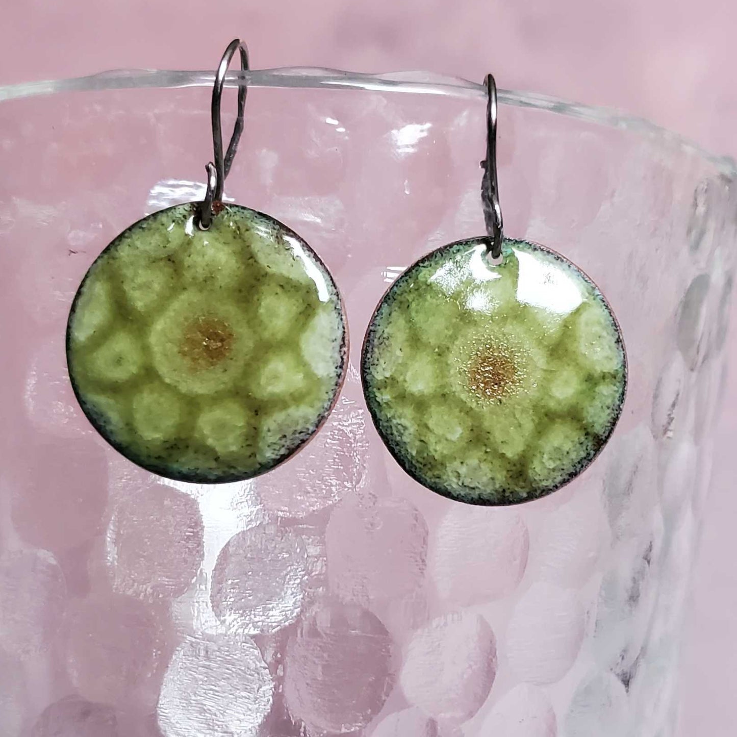 Green circular copper earrings on a glass surface with a pink background