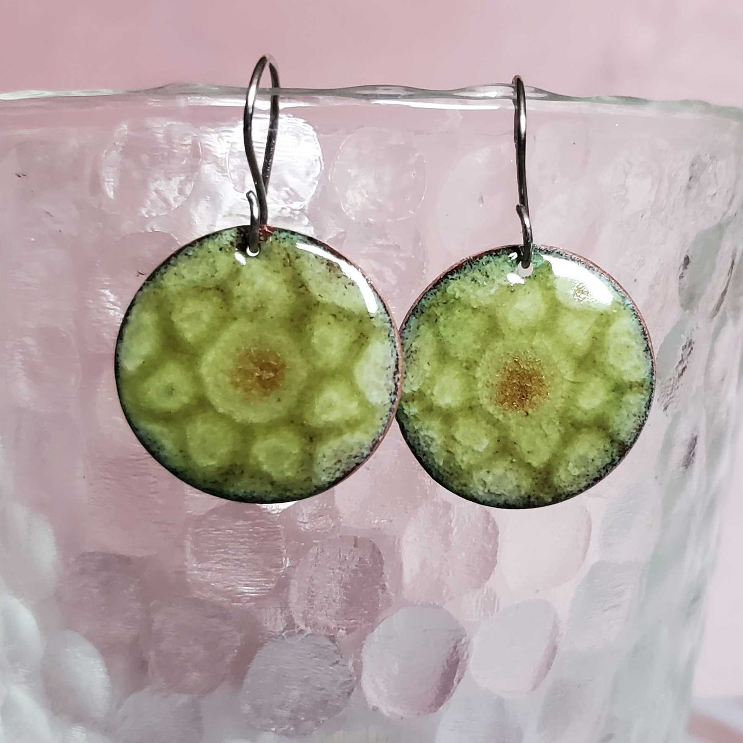 Green circular copper enamel earrings on a glass with a pink background