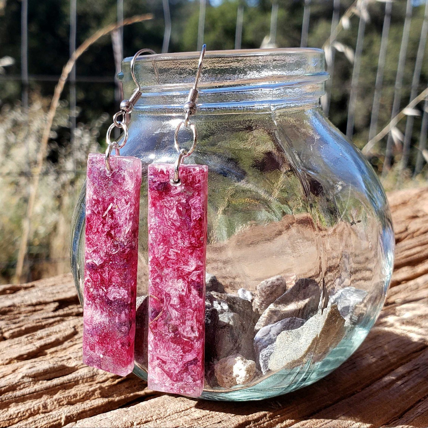 Pink dangle earrings with dried flowers
hanging from a glass jar outdoors.