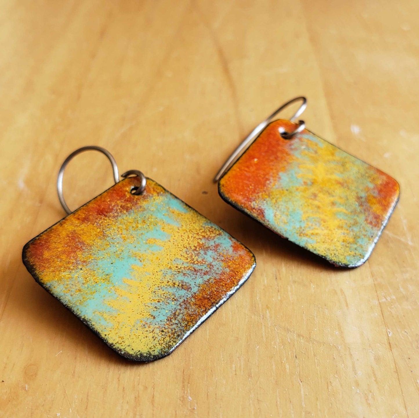 Pair of square copper earrings with multicolored patina on a wooden surface