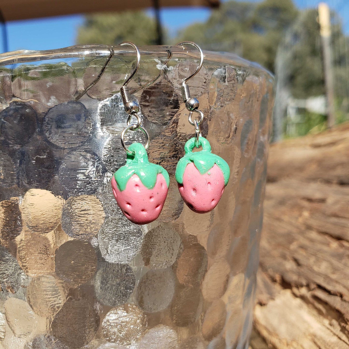 Pink and green strawberry-shaped earrings on a metallic surface with a natural background.