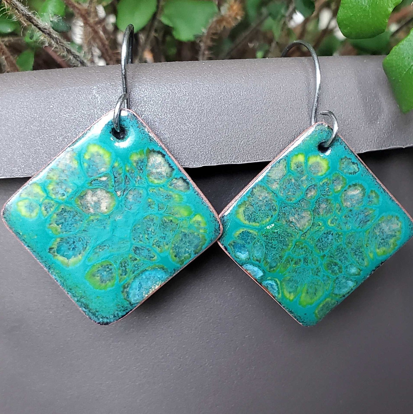 Teal and green patterned square copper earrings on a gray surface with a natural background.
