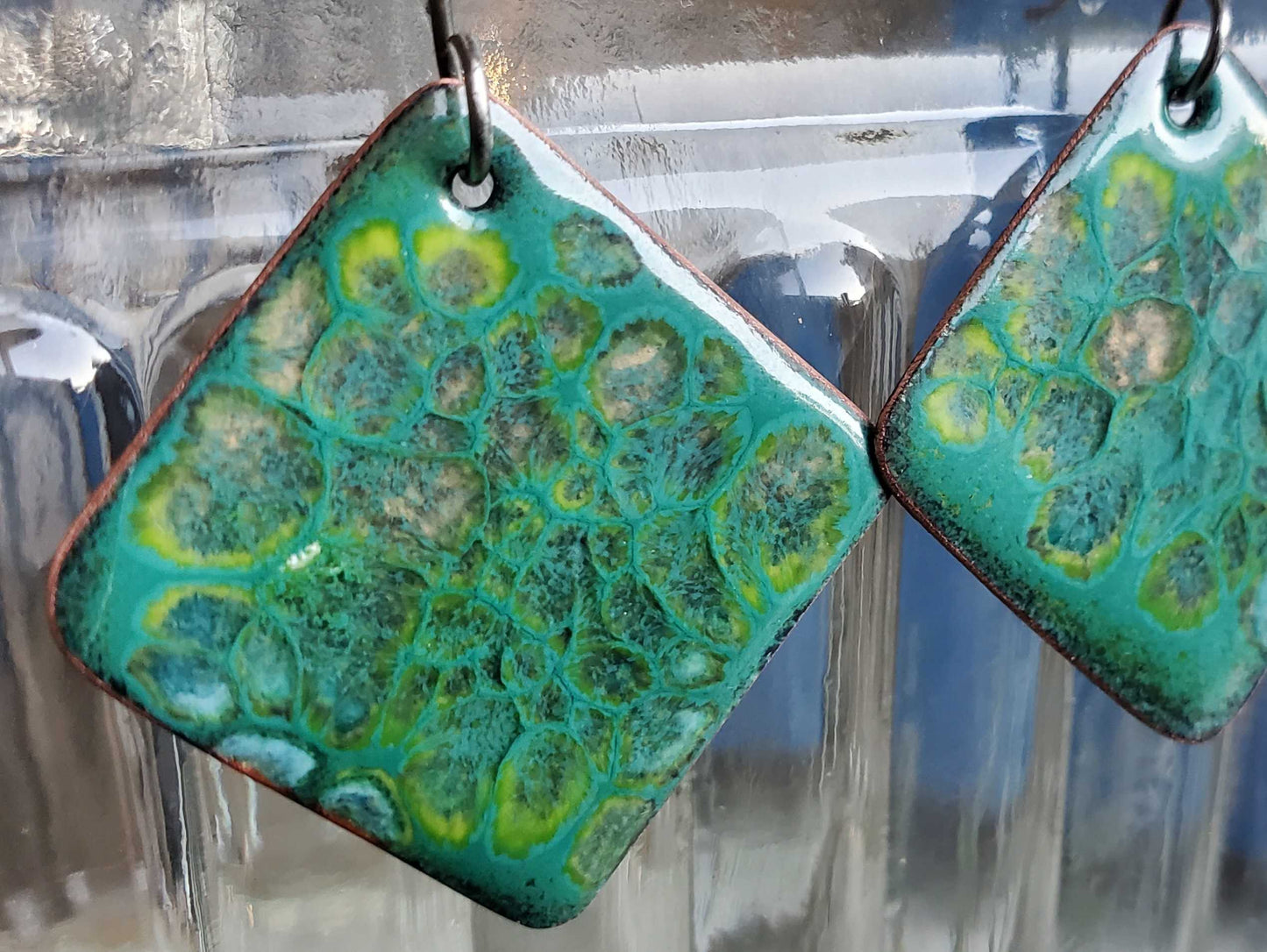 Green and blue patterned copper earrings hanging against a reflective glass surface.