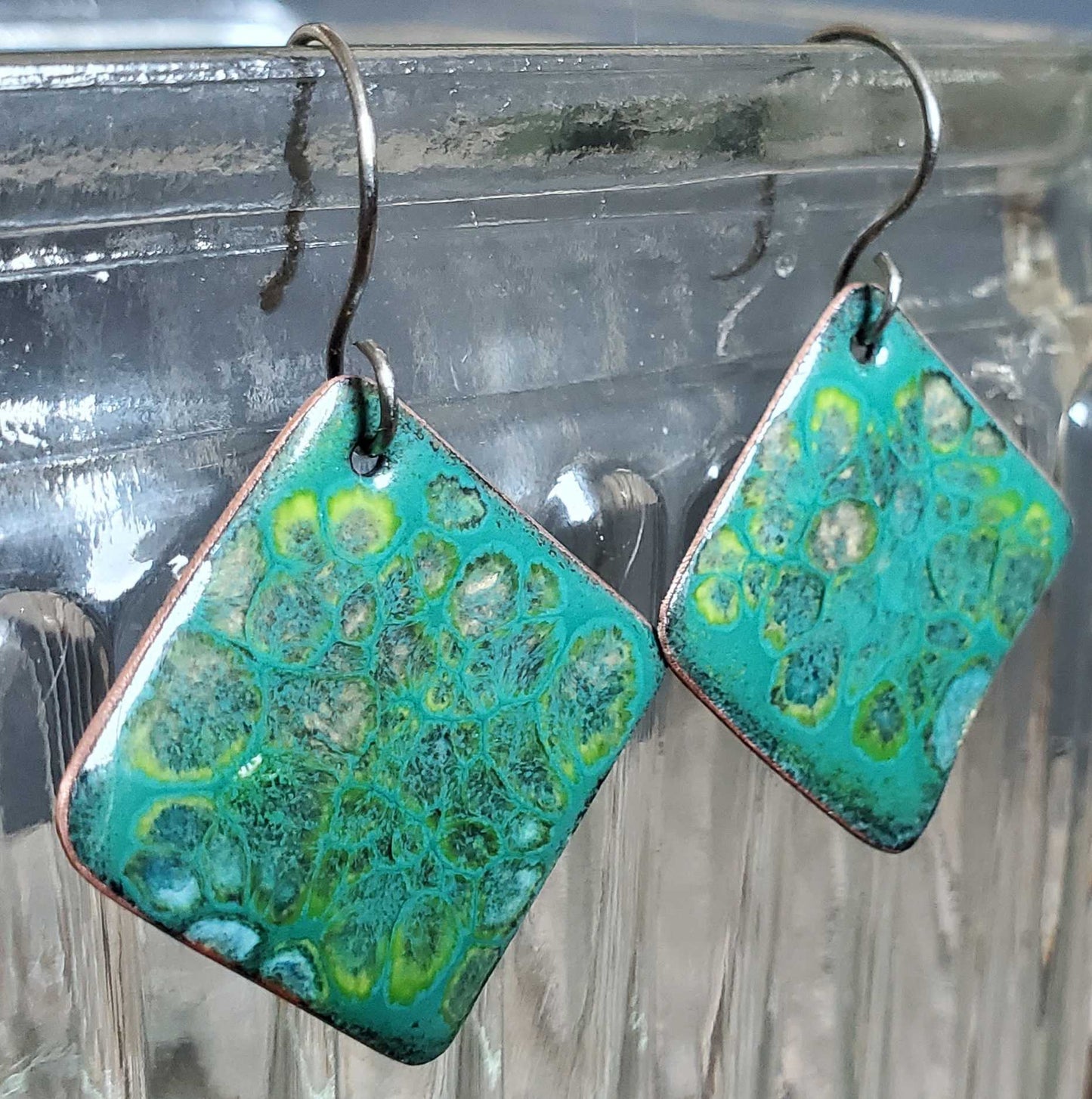 Pair of turquoise and green patterned copper earrings on a clear stand.