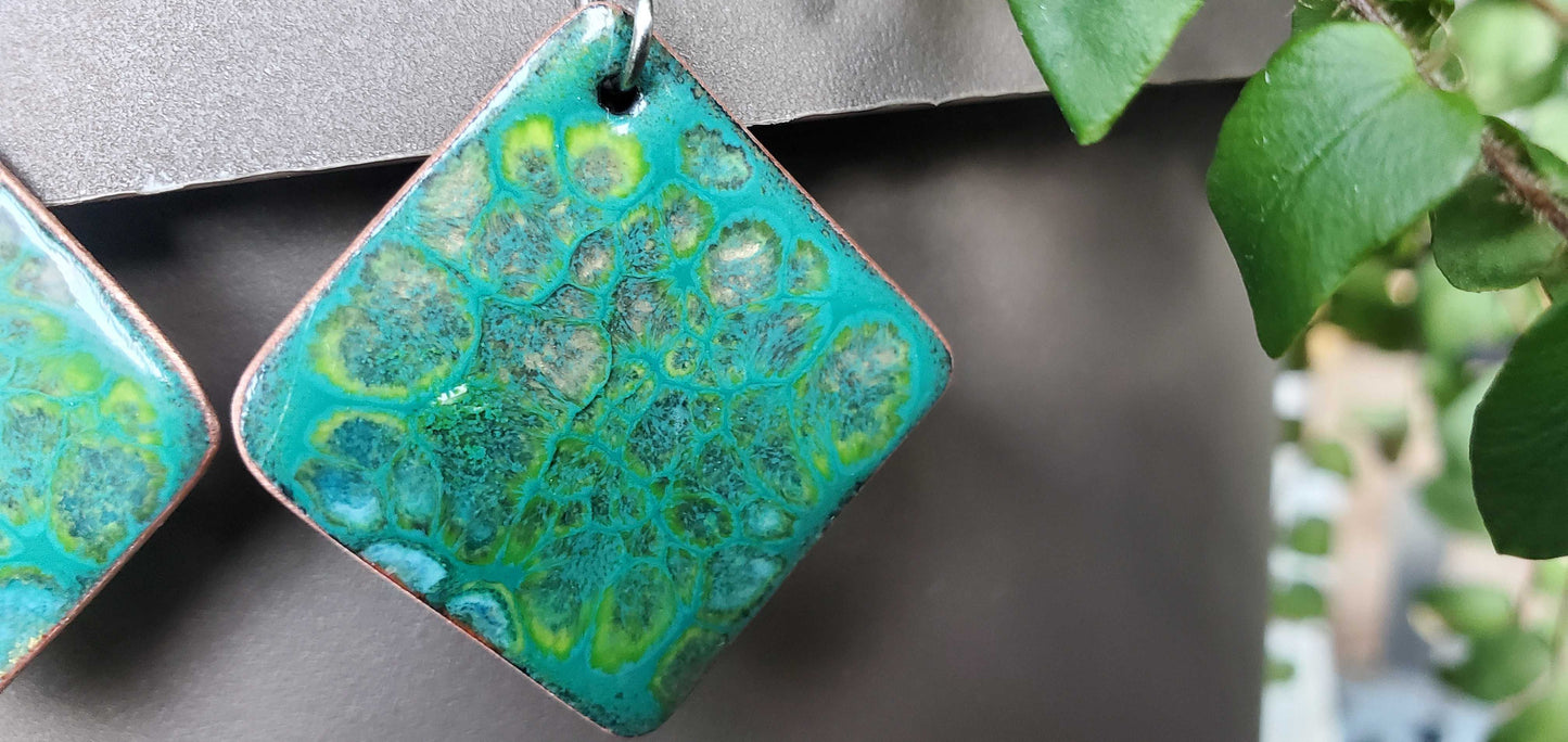 Teal and green marbled patterned copper earrings on a gray background with blurred green leaves.