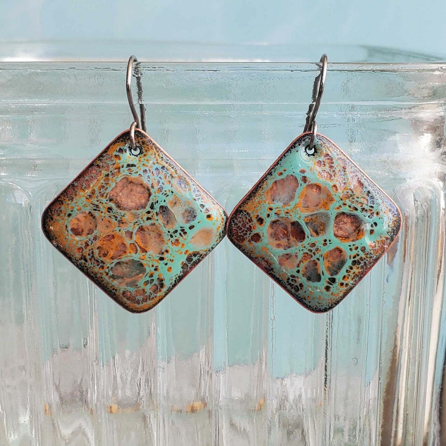 Pair of square copper earrings with crackle pattern hanging from a glass jar.