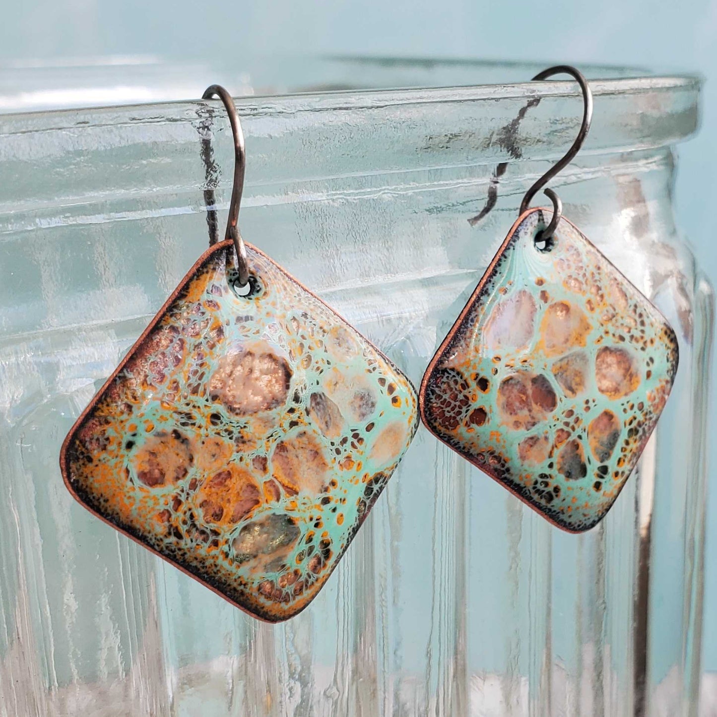Pair of square-shaped copper earrings with floral pattern hanging on a glass jar.