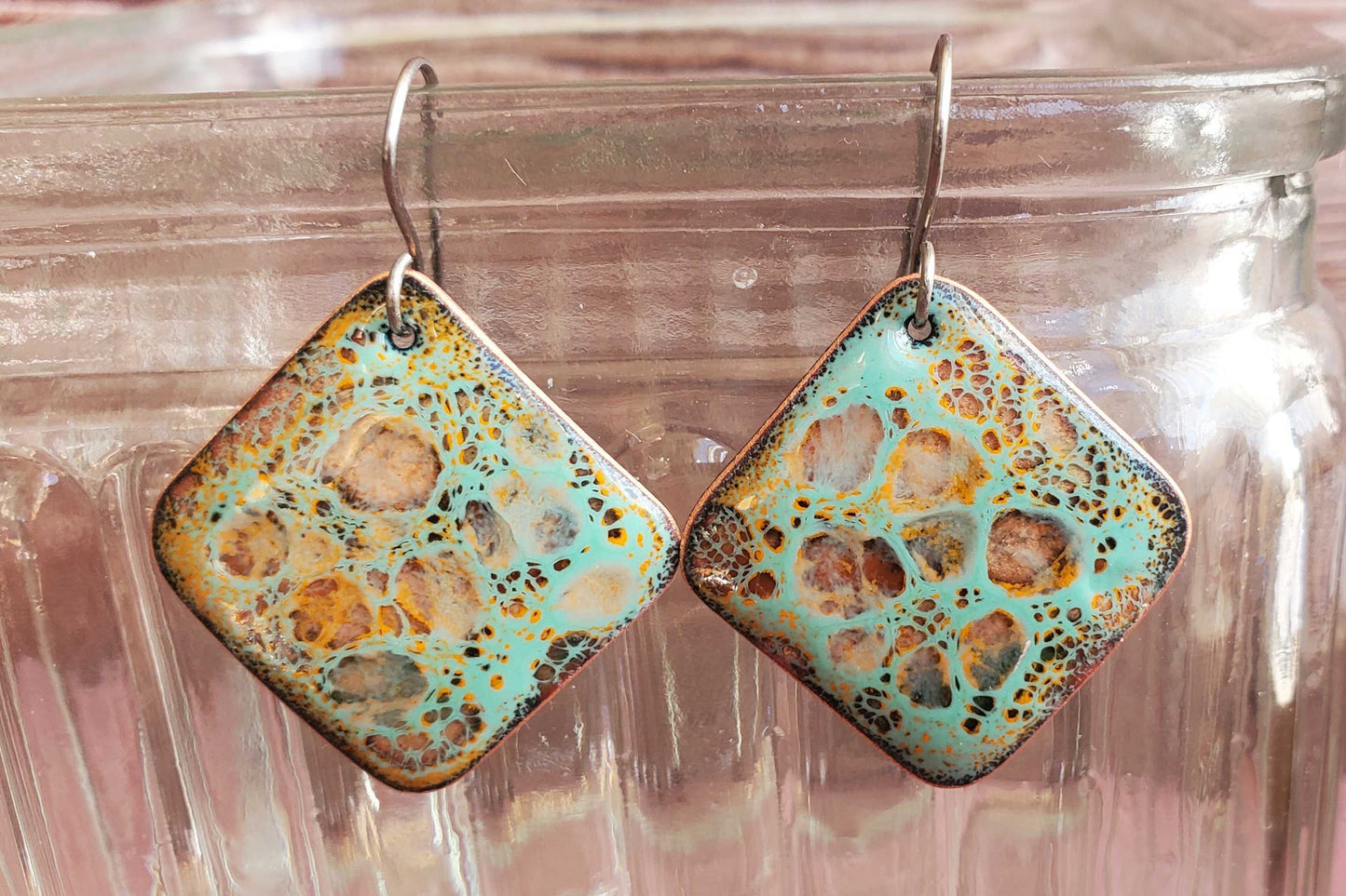 Square copper earrings with a textured, multicolored design hanging from a glass jar.
