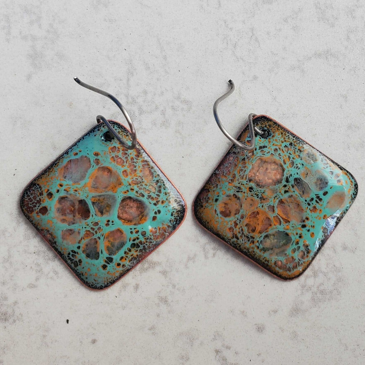 Pair of square copper earrings with a textured, multicolored surface on a light gray background
