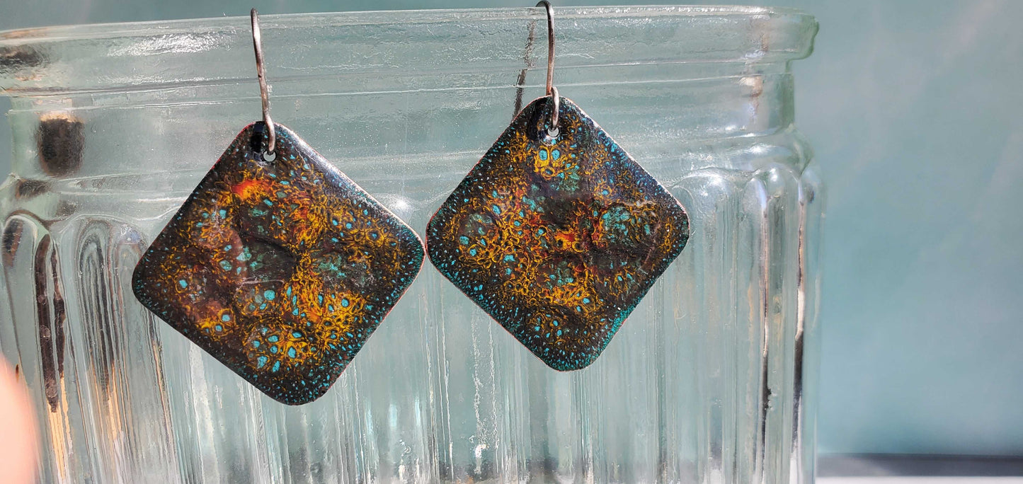 Square copper enamel earrings with a textured, multicolored design hanging from a glass container.