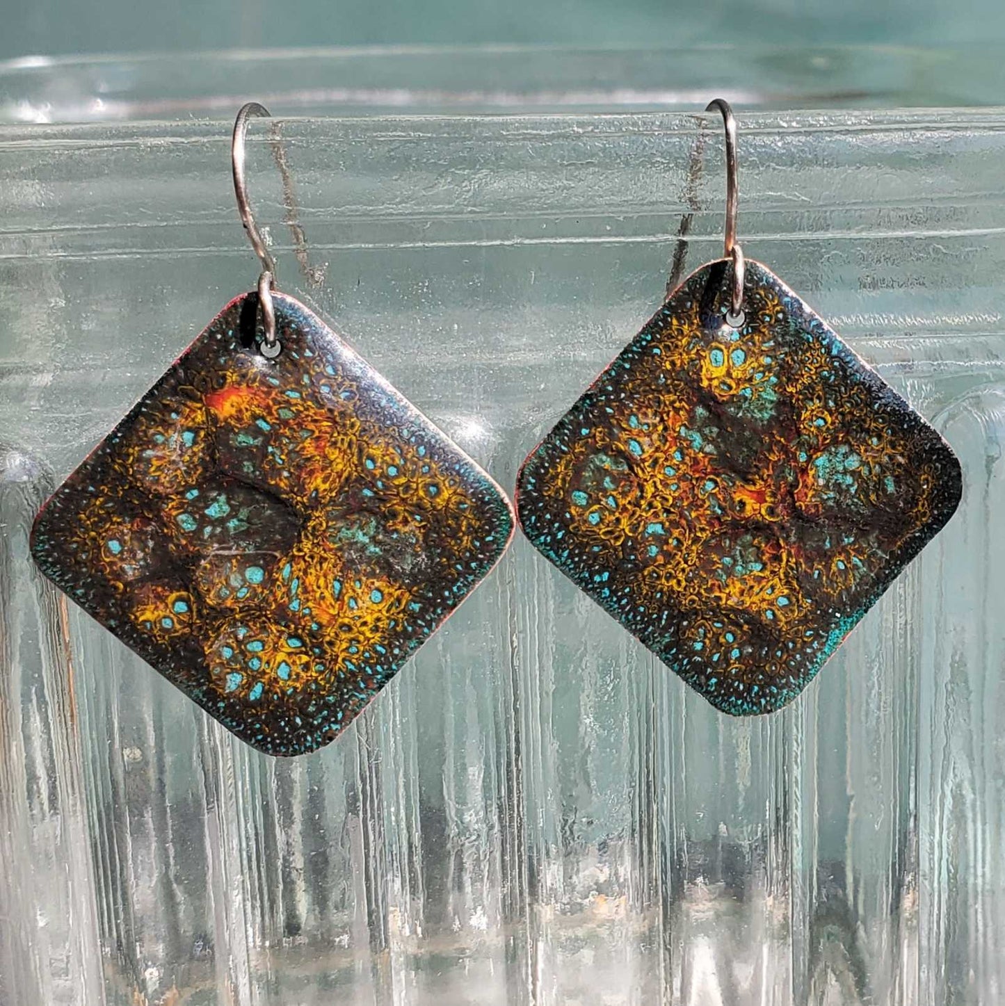 Pair of square copper earrings with a patterned design on a clear glass surface
