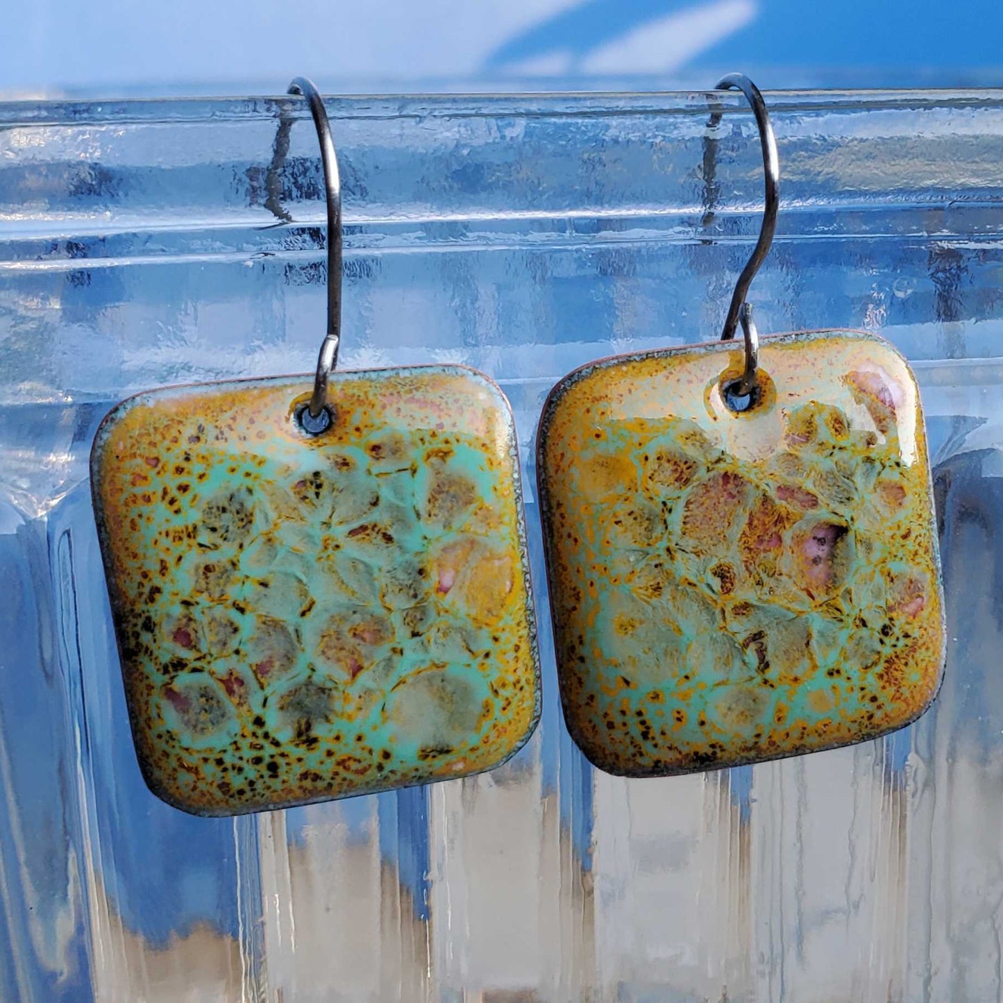 Pair of square copper and glass earrings with a textured surface on a clear stand.