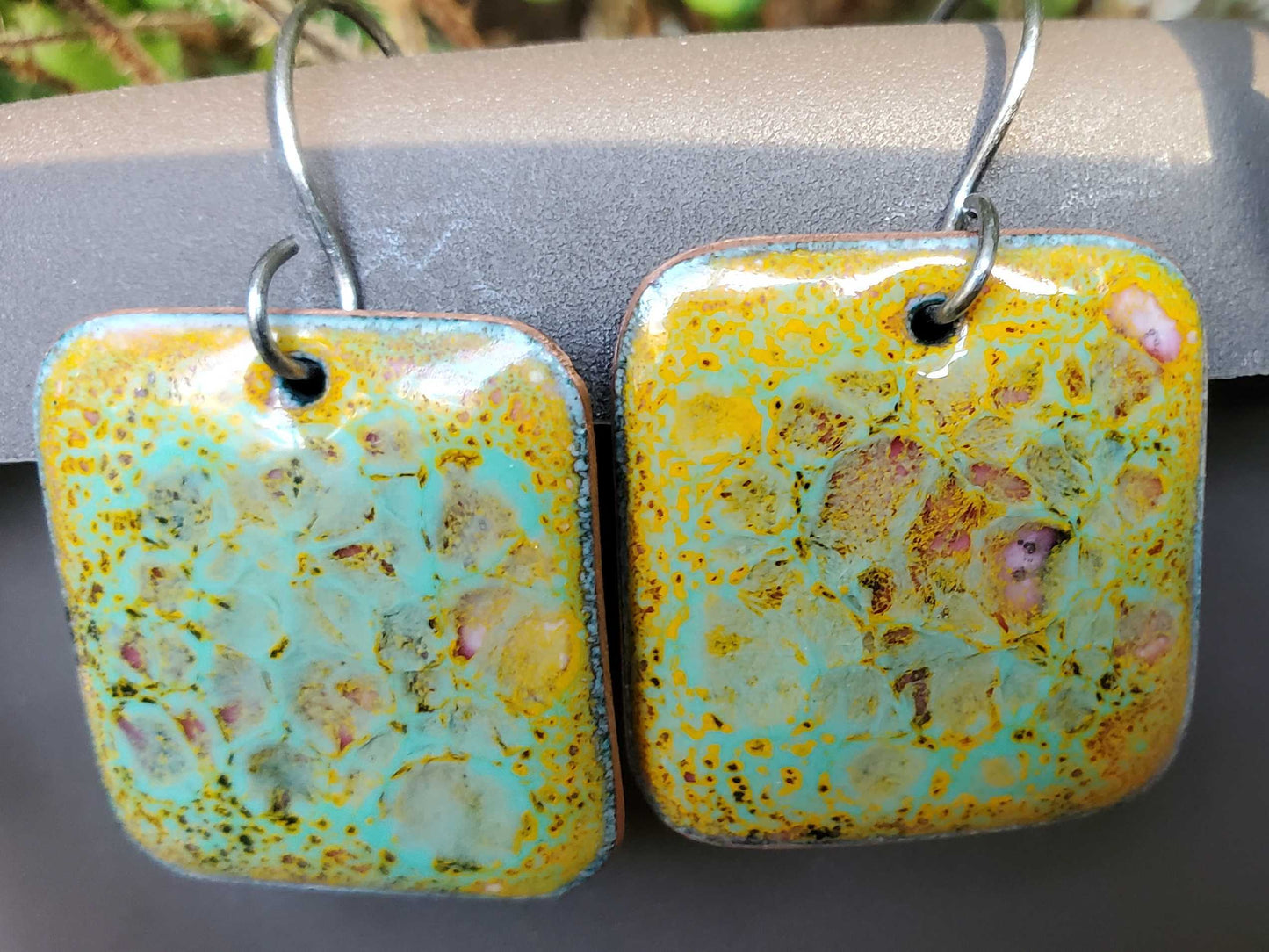 Rectangle copper and glass earrings with a multicolored glaze on a gray surface.