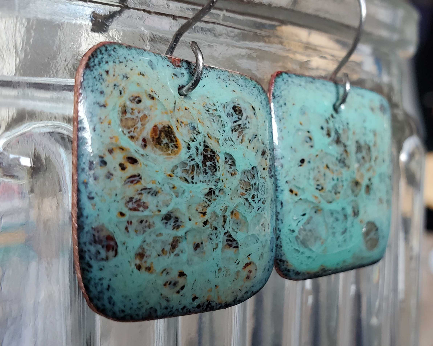 Turquoise copper and glass earrings with speckled pattern on a clear stand.