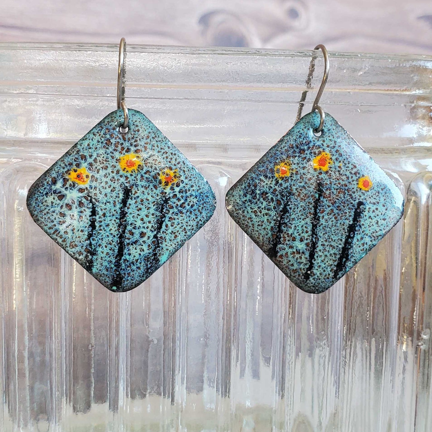 Pair of square copper enamel earrings with floral design on a glass surface