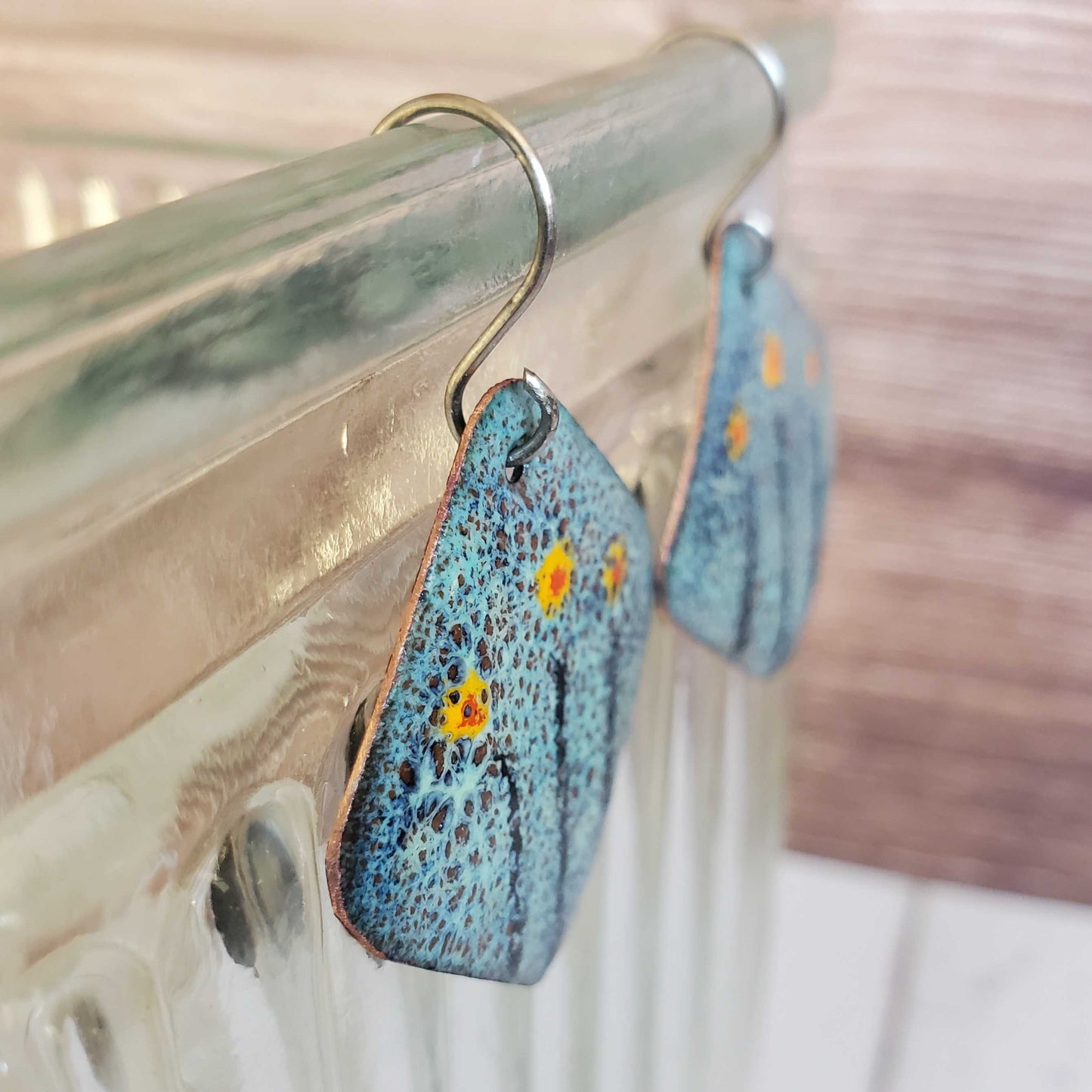 Blue copper enamel earrings with floral patterns hanging on a metallic surface.