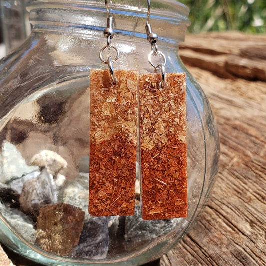 Rectangular earrings with a textured brown color made from dried leaves hanging in front of a glass jar with rocks