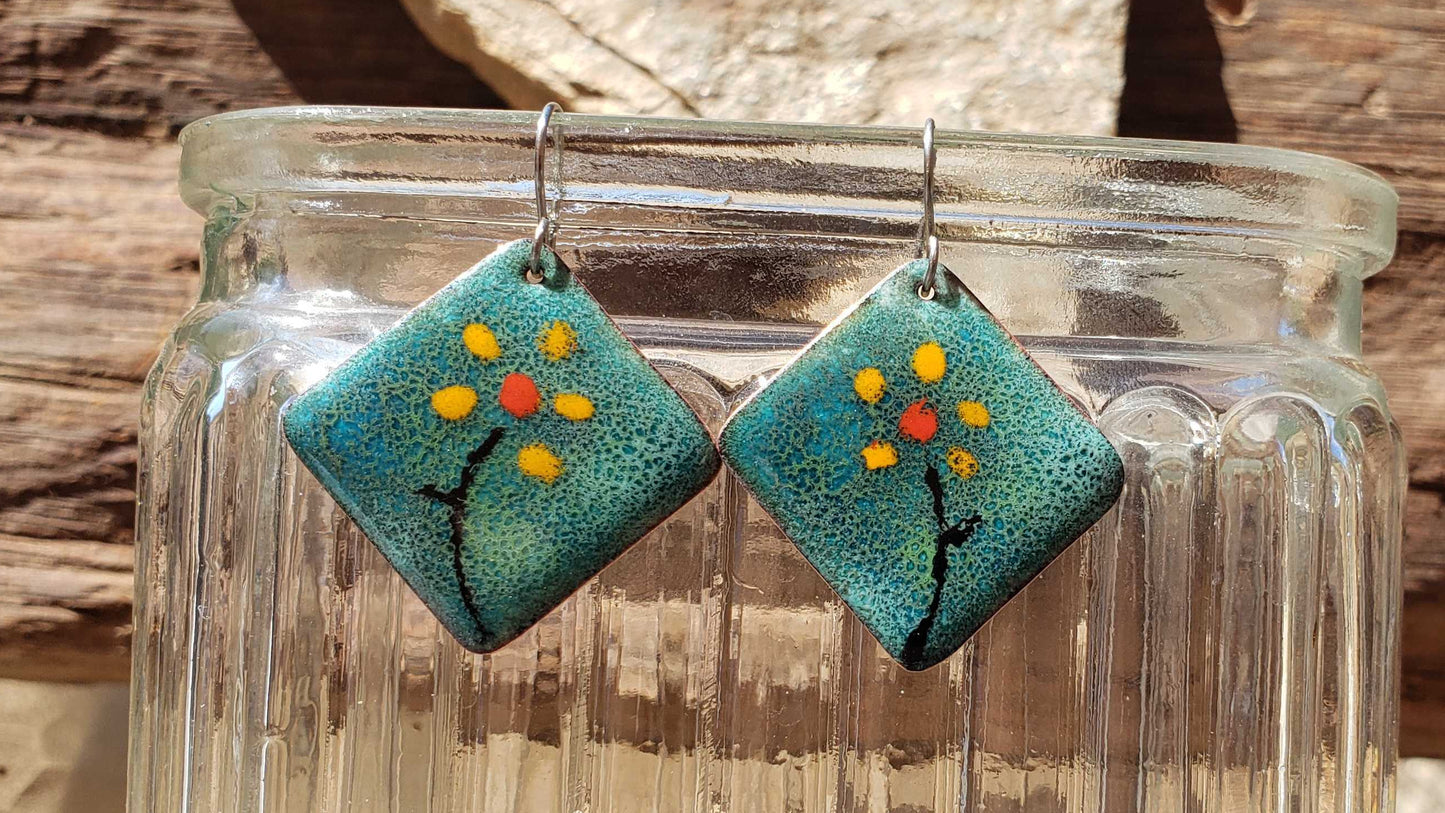 Pair of copper earrings with floral design hanging from a glass jar against a natural background.