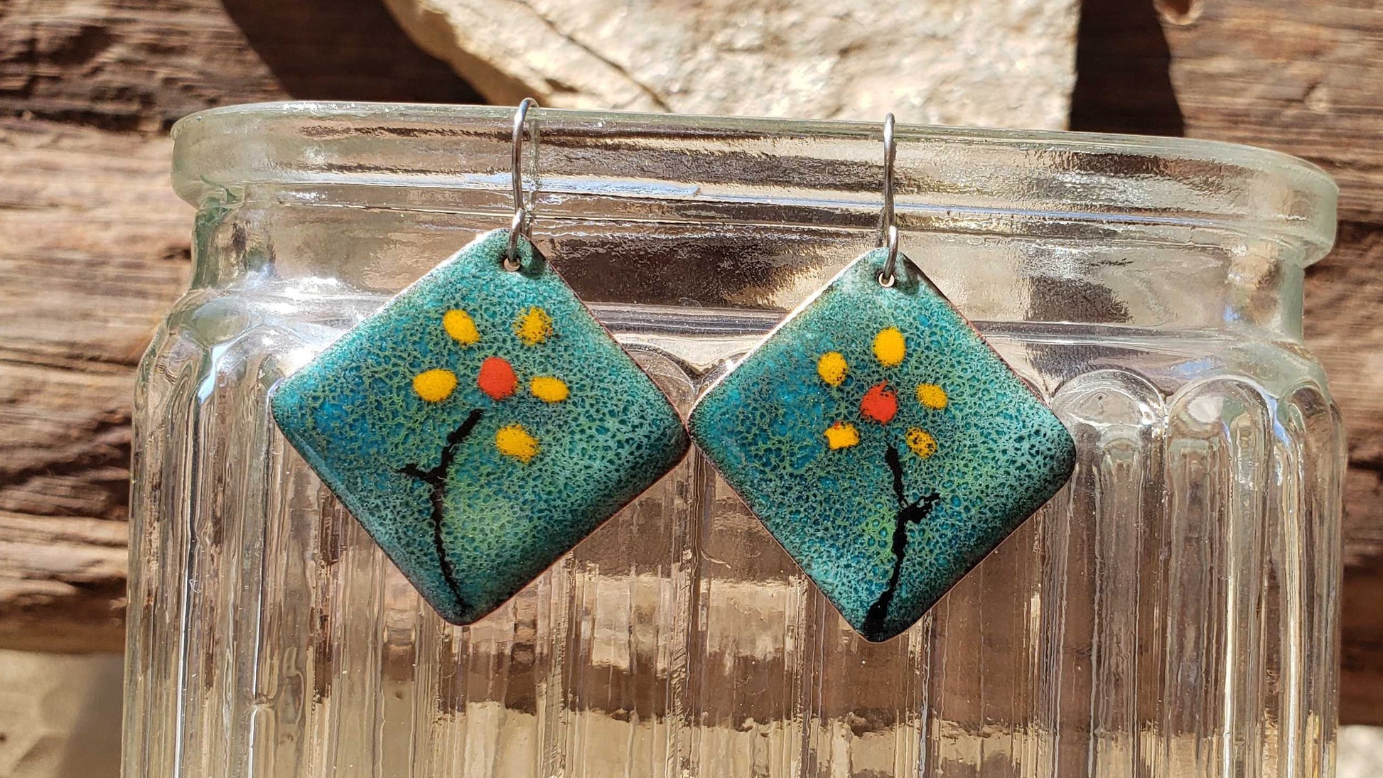 Pair of copper earrings with floral design hanging from a glass jar against a natural background.