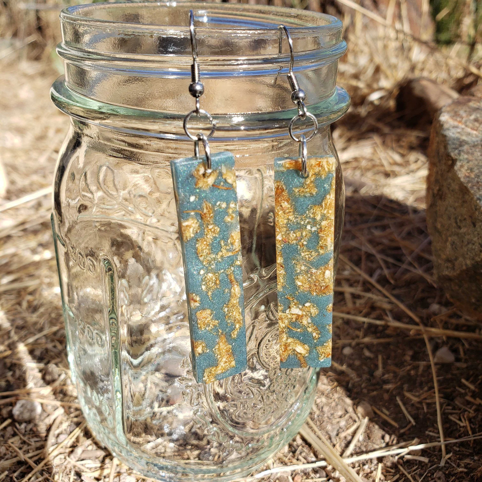 Pair of earrings with blue and gold design hanging from a glass jar on a natural background.