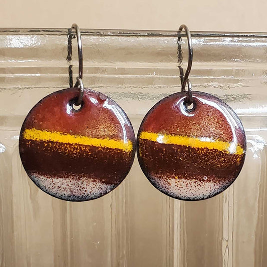 Round copper earrings with a brown and gold design hanging from a glass jar.