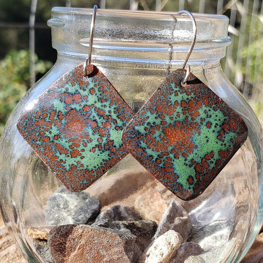 Square copper enamel earrings with green and brown patterns in a glass jar with stones