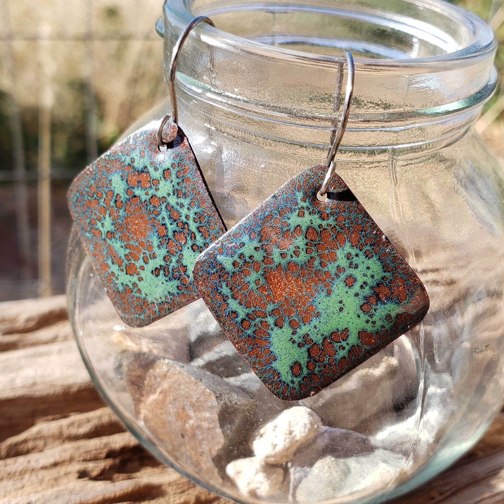 Square copper enamel earrings with green and brown pattern hanging from a glass jar.